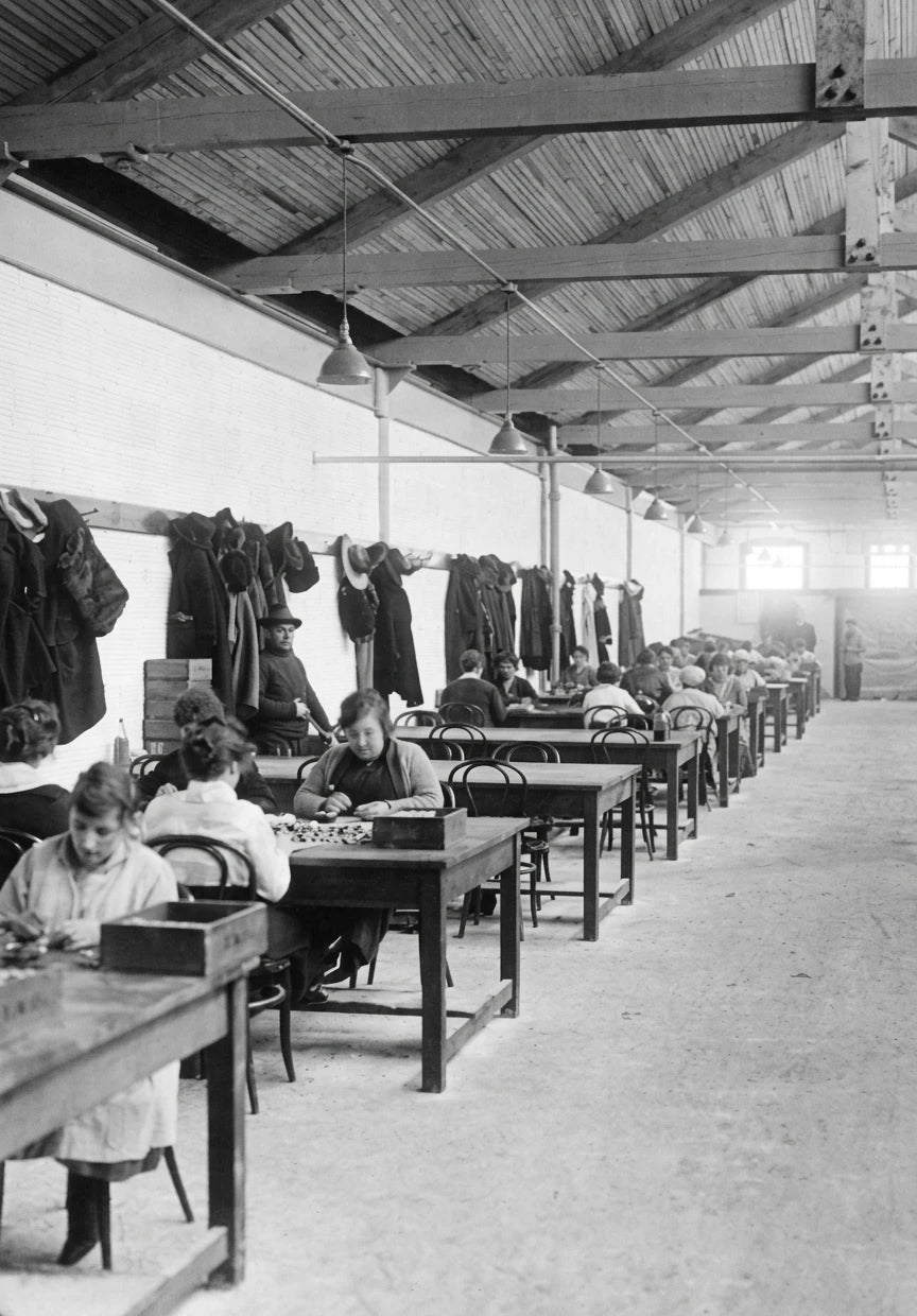 Vintage black and white photograph of a factory interior with workers at tables and hanging garments.